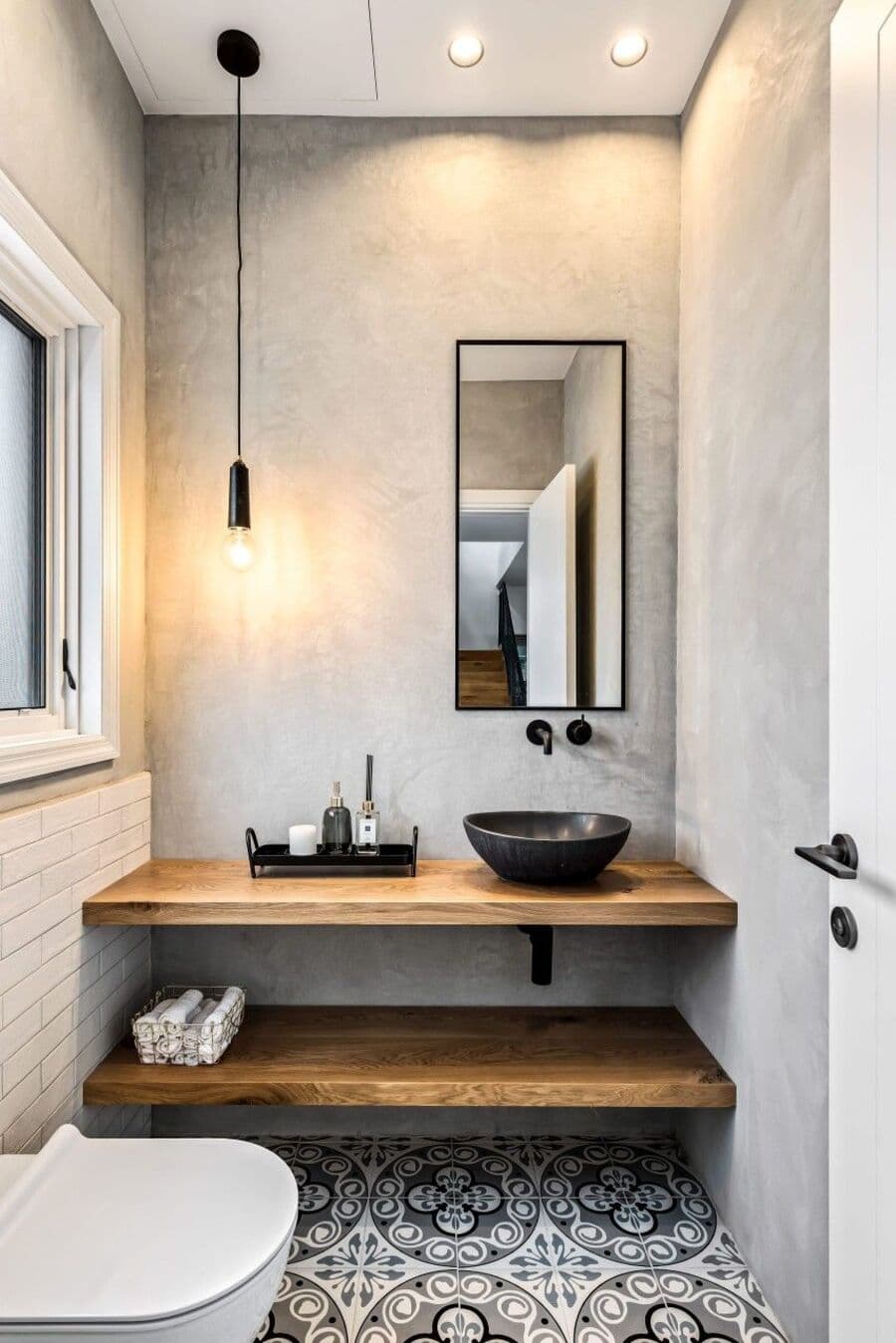 Baño rústico moderno con baldosas hidráulicas y muro de cerámica blanca y pintura gris. Vanitorio tipo repisa de madera con lavamanos redondo negro. Lámpara de estilo industrial, con ampolleta a la vista.