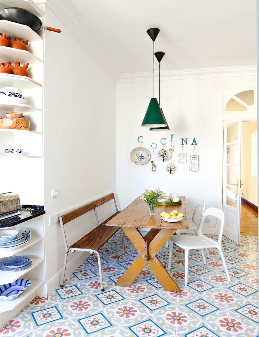 Cocina estilo ecléctico romántico con comedor de diario. Mesa estilo picnic de madera con una banqueta de madera y sillas blancas. Piso de baldosas hidráulicas en tonos azul y tierra. Sobre la mesa hay dos lámparas colgantes de color verde. Muros blancos