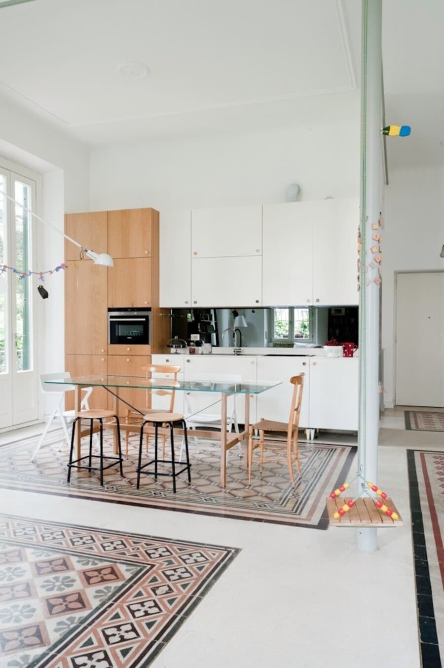 Cocina abierta con comedor integrado, de estilo moderno. Comedor con mesa de vidrio y sillas y pisos de madera. Bajo la mesa, hay un sector de baldosas hidráulicas demarcando el comedor. El resto del piso es de cerámica blanca.