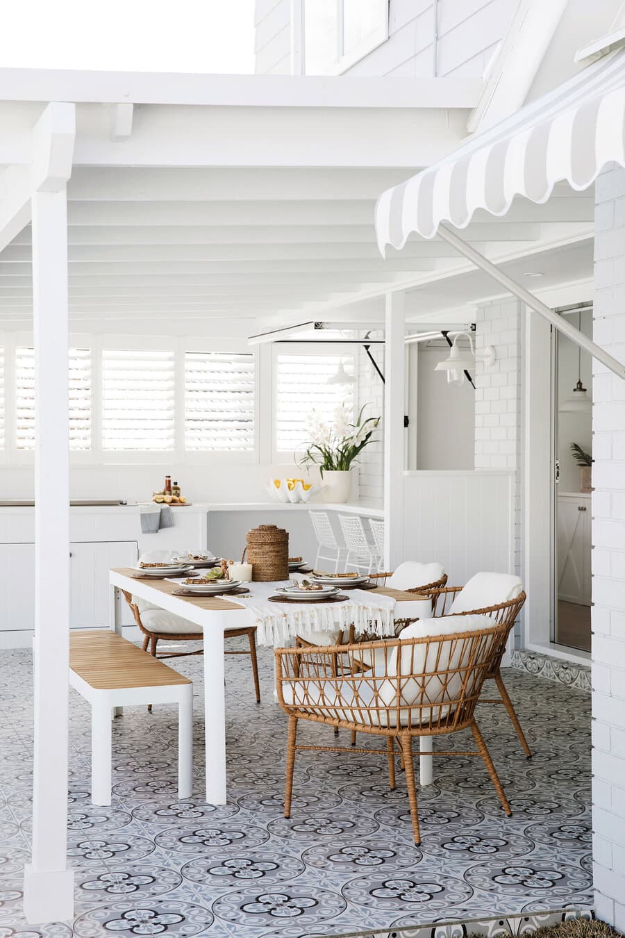 Terraza techada estilo playero. Muros y techo blanco y piso de baldosas hidráulicas color gris y negro. Mesa de comedor de metal blanco y base de madera, banqueta y sillas de mimbre con cojines blancos. 