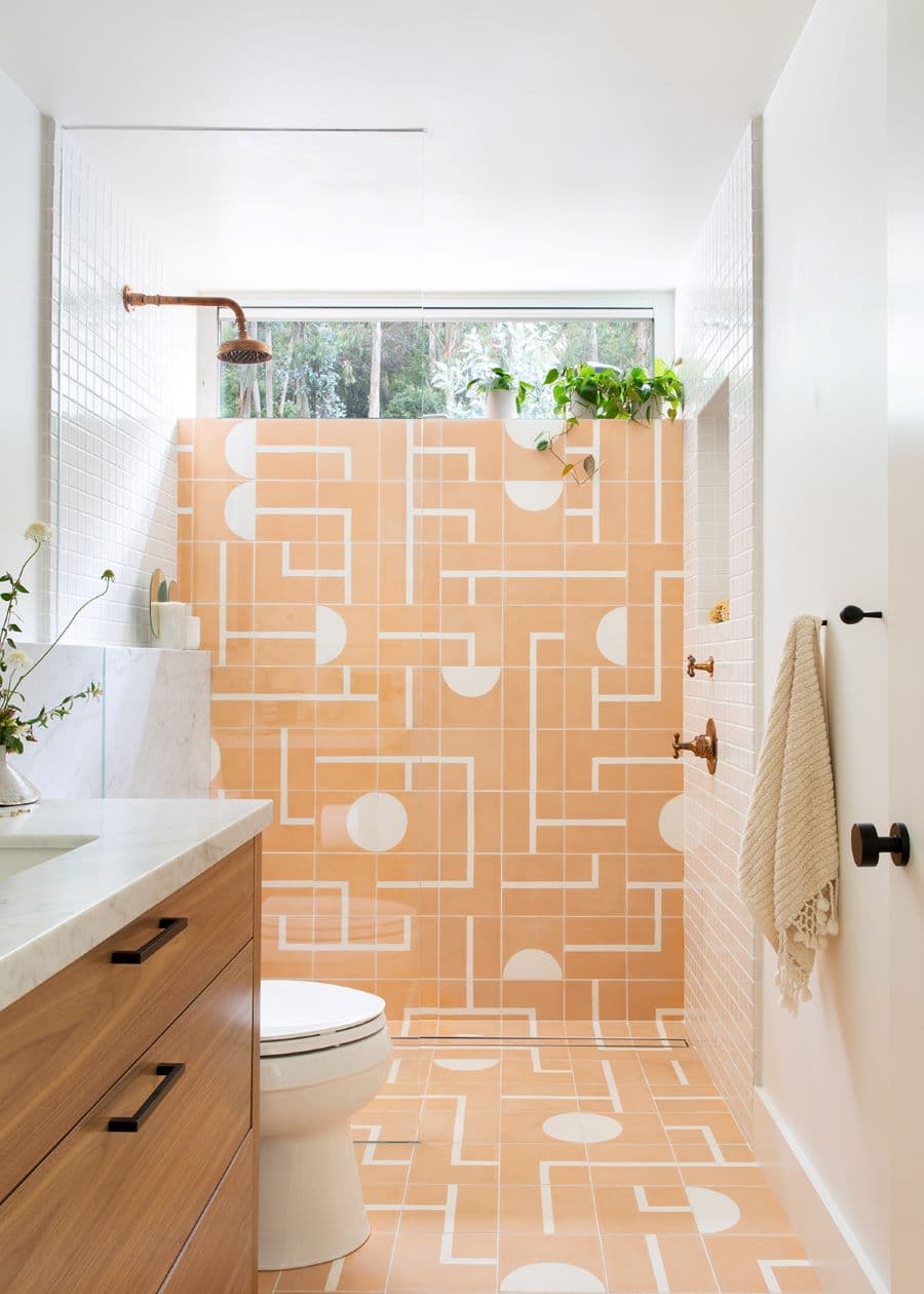 Baño moderno, con azulejos geométricos en color damasco y blanco. También tiene azulejos cuadrados pequeños y un muro pintado blanco. Mampara de vidrio y grifería de cobre. Vanitorio de madera con base de mármol.