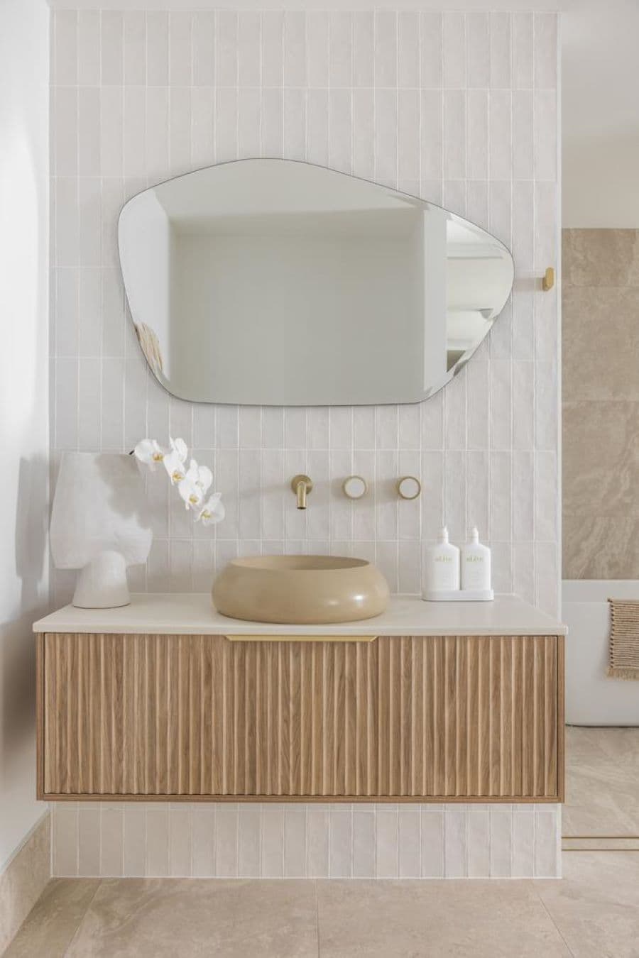 Baño moderno, con azulejos blancos, rectangulares. Vanitorio flotante con puertas de madera y un tirador largo de color dorado. Lavamanos redondo, sobrepuesto, color beige oscuro. Grifería empotrada en el muro. Gran espejo colgado, de forma irregular.