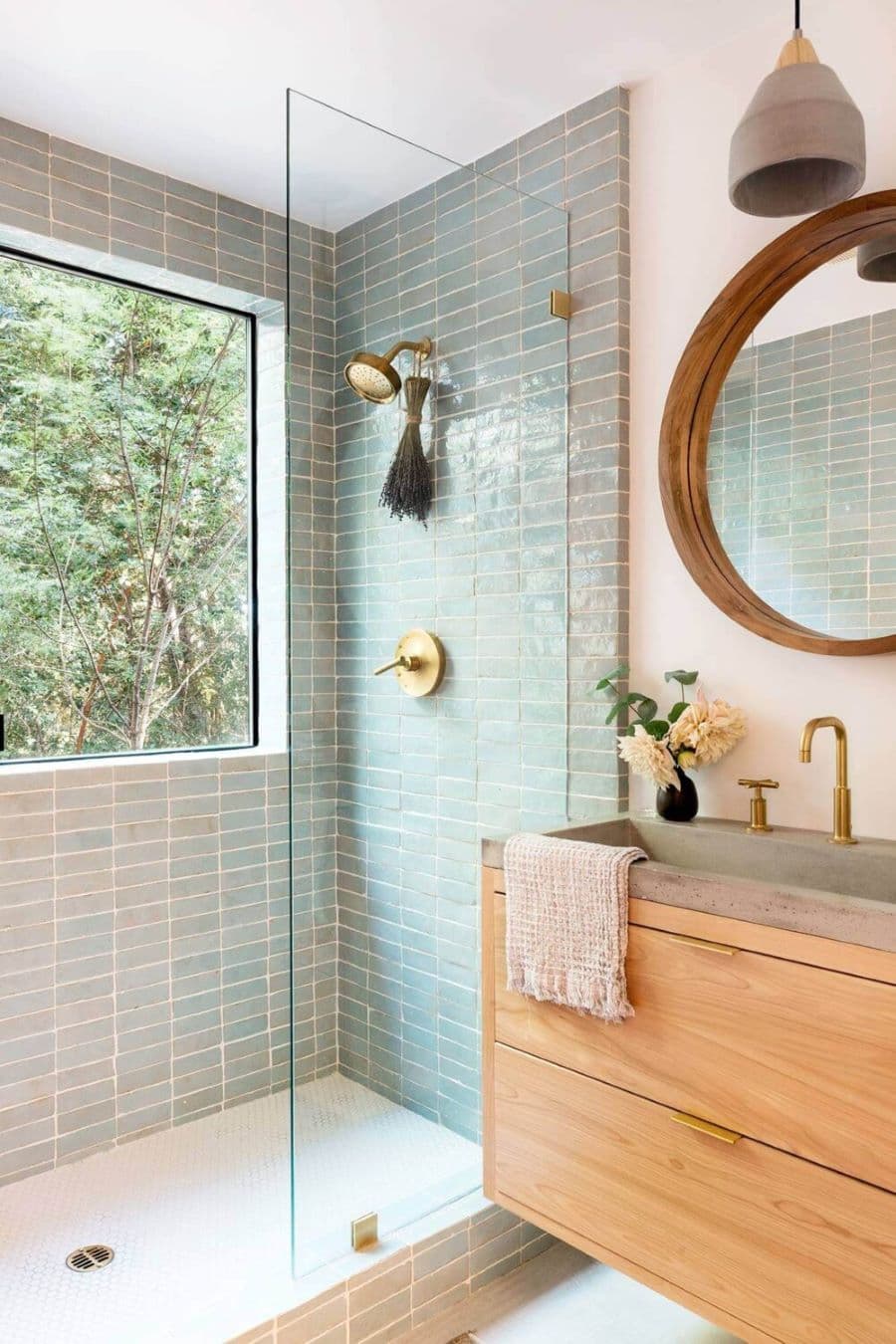 Baño con ducha con azulejos rectangulares color celeste oscuro. Grifería dorada. Mampara fija de vidrio templado. Vanitorio flotante, de madera y base de concreto. Espejo redondo con marco de madera y lámpara colgante de estilo industrial.