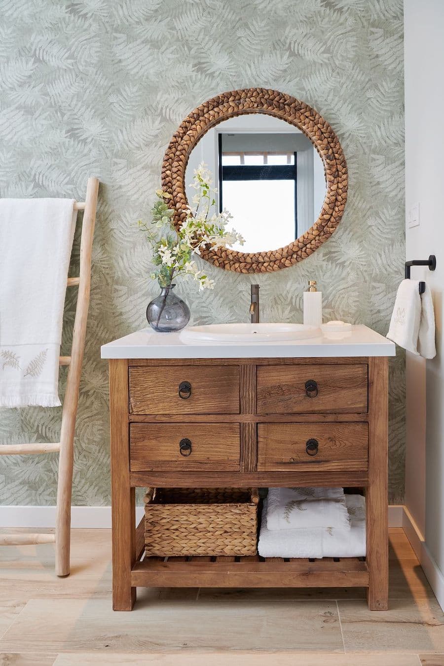 Baño rústico moderno con papel mural con hojas verdes. Mueble vanitorio de madera con 4 cajones y base blanca. Espejo redondo con marco de mimbre. 