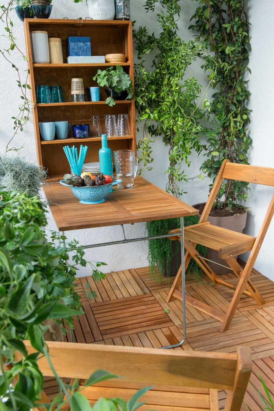 Balcón con mesa plegable de madera. La mesa está abierta y en la parte del muro tiene dos repisas que tiene vasos y vajilla celeste. Hay una silla de madera plegable. El piso es de madera y los muros son blancos. Muchas plantas en maceteros