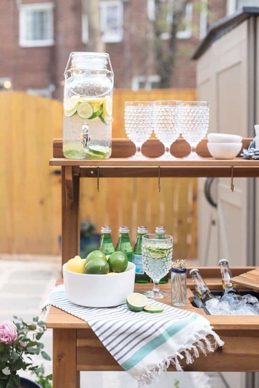 Repisa de madera con jarro dispensador, de vidrio, con agua con limón. Tres copas con diseño y dos bowls pequeños blancos. En la parte de abajo hay un bowl con limones, hielera con botellas, una copa con líquido, un paño de cocina.