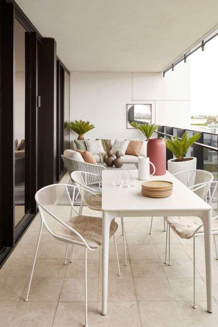 Gran terraza blanca, con baranda negra de metal. Tiene un área con un comedor, con mesa blanca y 4 sillas blancas con cojines. Atrás se ve otro sector de living con un sillón y dos sofás blancos con cojines de colores verde y tierra. El piso es de cerámica