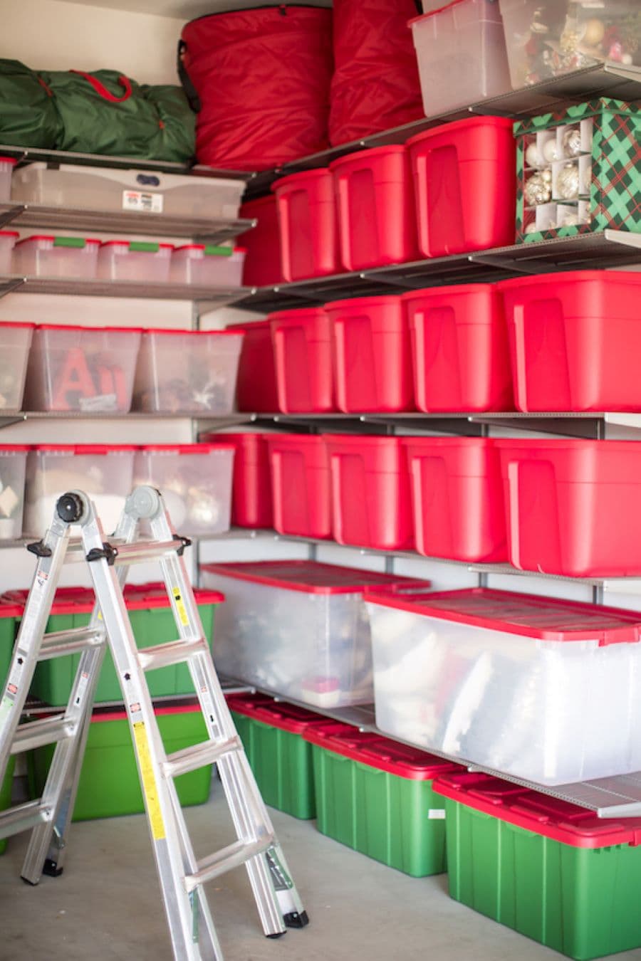 Bodega con cajas plásticas transparentes, rojas y verdes, que guardan adornos de navidad. También hay bolsas. Las cajas están sobre repisas flotantes pegadas a la pared. Hay una escalera en A, abierta sobre el suelo