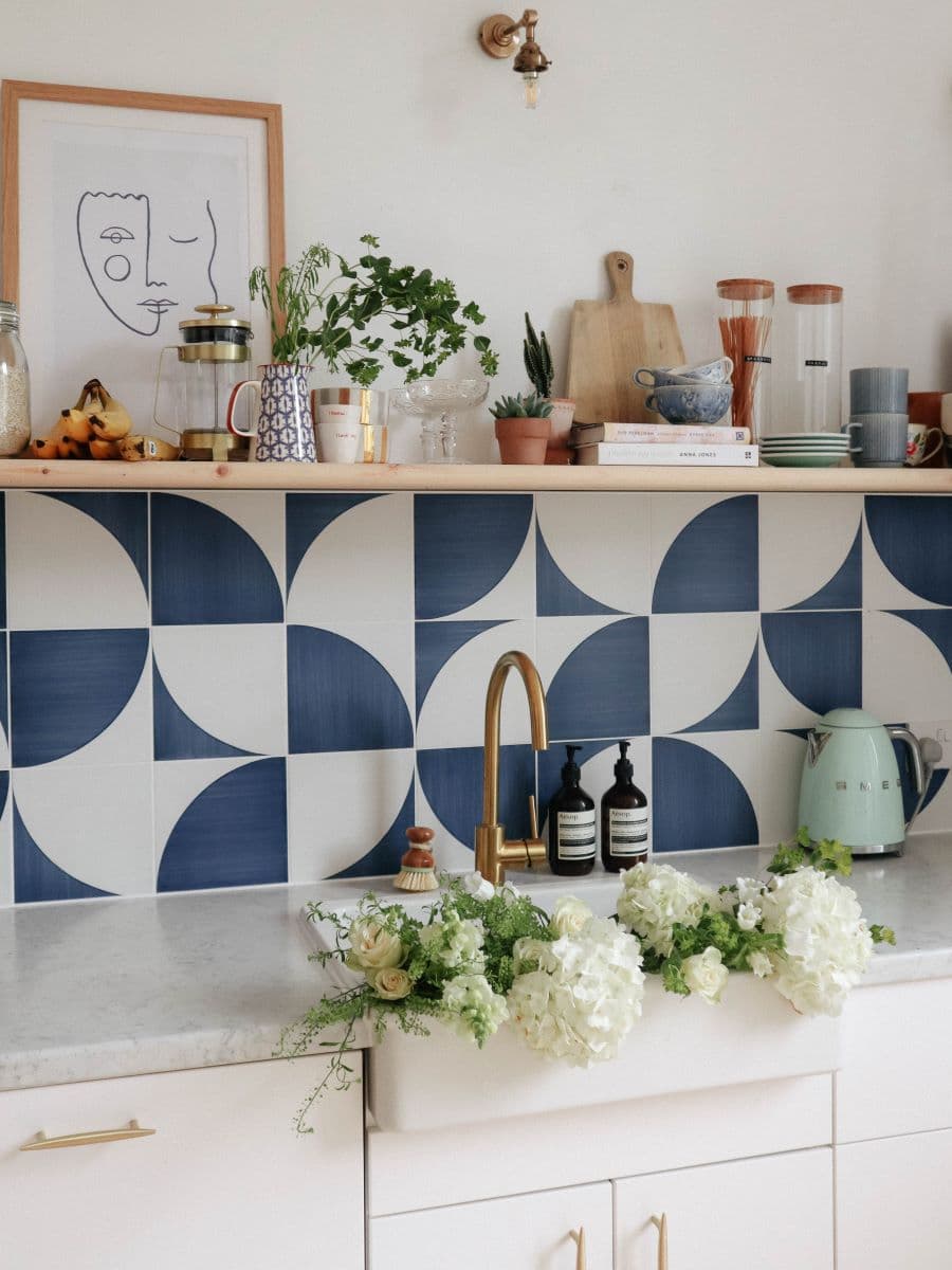 Cocina luminosa con cerámicos de patrón geométrico, blanco y azul, en la parte baja del muro. Sobre ellos hay una repisa flotante de madera con adornos, plantas y cuadros. El mesón de la cocina es de mármol y tiene un lavaplatos blanco con grifería dorada