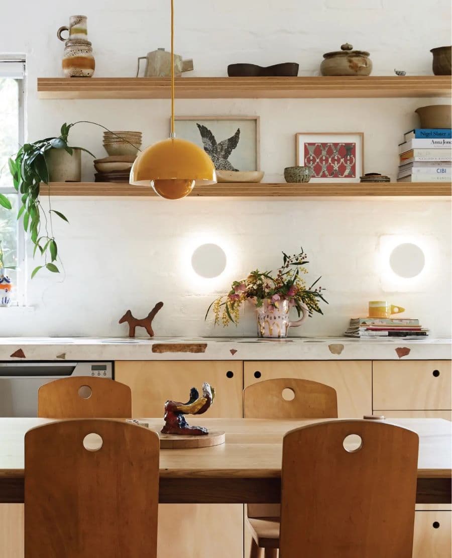 Cocina de estilo mid century con muebles de madera y dos repisas flotantes con cuadros, adornos y libros. Mesa de comedor con sillas de madera. Lámpara blanca colgante, de color amarillo. En el muro hay dos focos led, redondos.