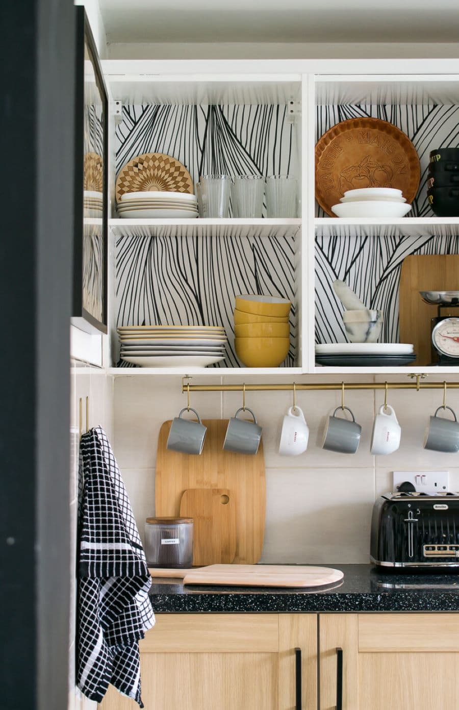 Muebles de cocina superiores abiertos, con fondo de papel mural blanco con negro. Vajilla blanca y de madera a la vista. Tazas colgadas bajo el mueble con pequeños cáncamos, Mueble con puertas de madera y mesón de cuarzo negro.