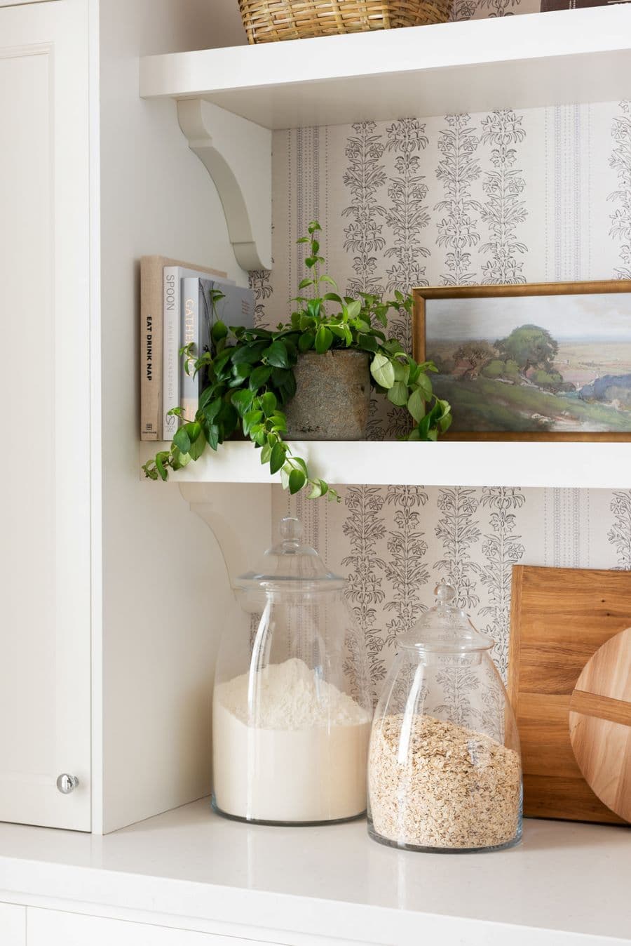 Detalle de mesón de cocina blanco con dos frascos de vidrio con avena y harina. Junto a ellos hay tablas de picar. Repisa flotante blanca con libros, planta y un cuadro. El muro tiene papel mural con pequeñas enredaderas negra.