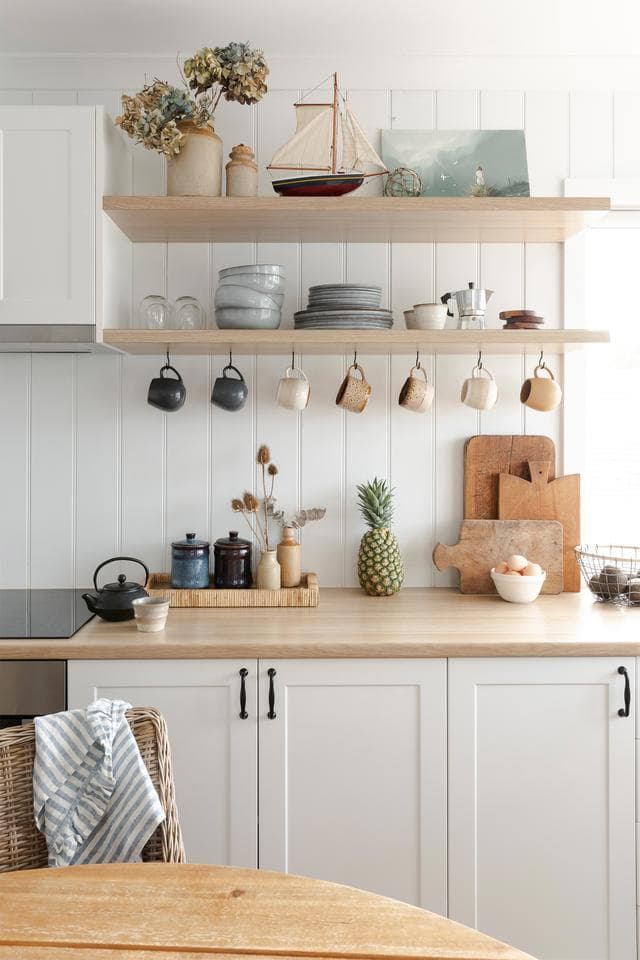 Cocina de estilo clásico con muebles de madera blanca y base de madera, Dos repisas flotantes con vajilla. Sobre el mesón hay una bandeja con utensilios, tablas de picar y una piña. Muro de revestimiento de madera blanca.