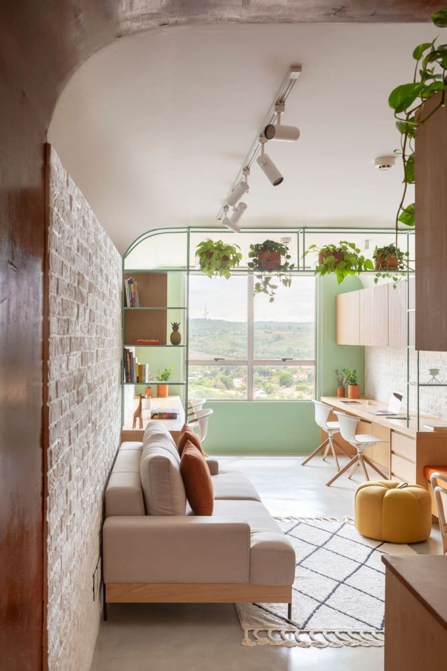 Departamento pequeño con muro de ladrillo, piso blanco. Gran ventana con un marco donde hay adornos, libros y plantas. Sillón gris y alfombra blanca con líneas negras. Pouff amarillo, escritorio a medida, de madera, con dos sillas blancas.