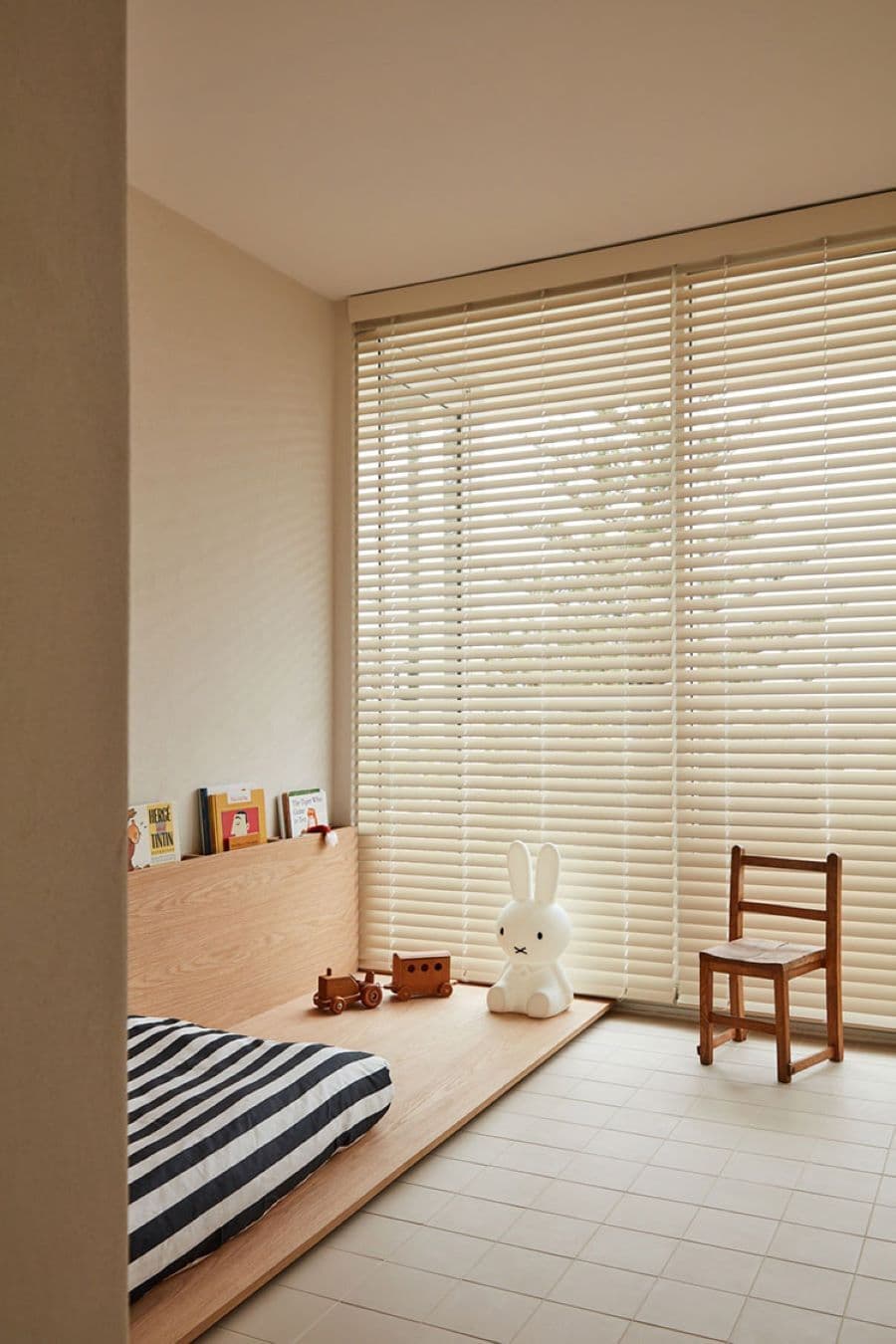 Dormitorio infantil con un gran ventanal con persianas. El piso es de cerámicas cuadradas de color beige. La cama está en el suelo sobre una base de madera. Hay una pequeña silla al costado y un conejo decorativo.