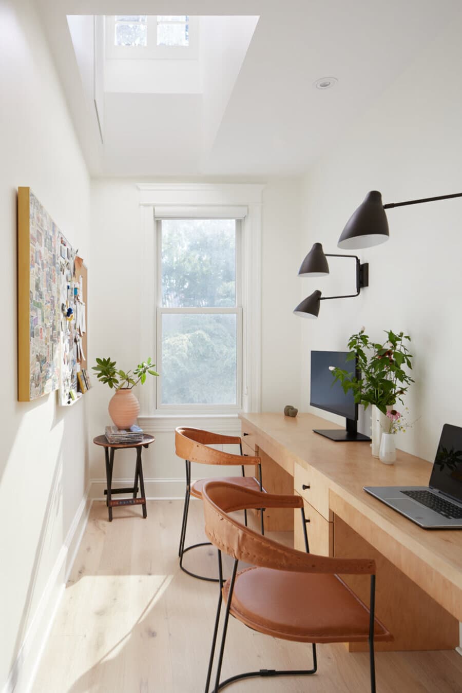 Oficina pequeña con un escritorio doble de madera. Dos sillas de metal y cuero. Los muros son blancas y el piso es de madera clara. Hay una gran ventana y una mesa de apoyo negra con una planta. Dos lámparas de muro estilo industrial