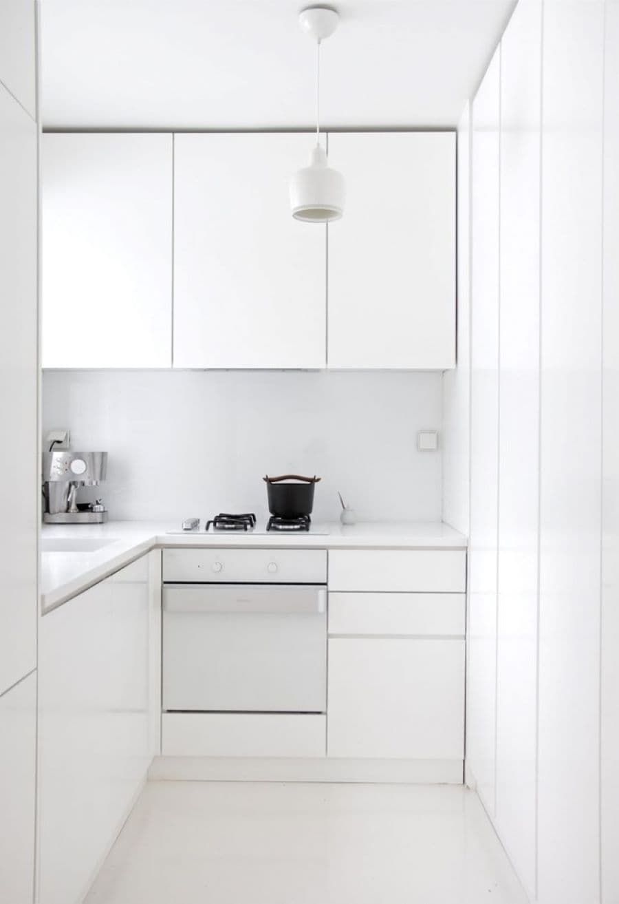 Cocina pequeña minimalista, con paredes y muebles color blanco. Cafetera cromada en una esquina y pequeños electrodomésticos negros.