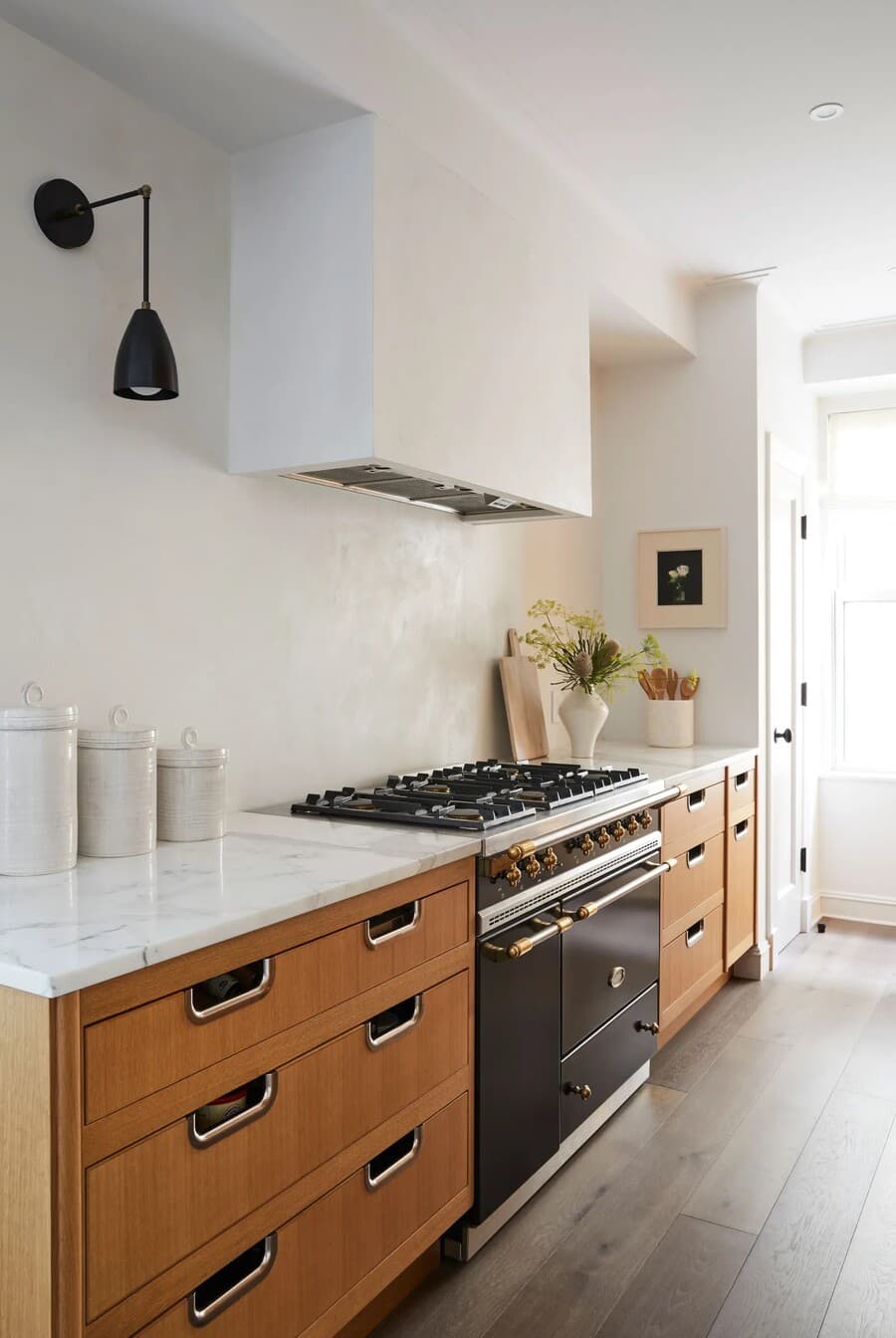 Cocina con muros blancos texturados, muebles de madera rojiza y mesones de mármol. Gran cocina a gas con una campaña con revestimiento blanco. Sobre el mesón hay tres contenedores de cerámica, tablas de madera, un florero y un recipiente con cucharas 