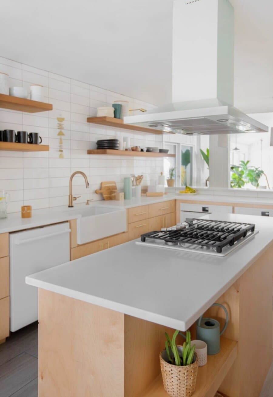 Cocina abierta, con muros cn cerámicos blancos. Muebles de madera y mesón de cuarzo blanco. Cocina a gas en una isla y sobre ella una gran campana colgada del techo. 4 repisas flotantes de madera con vajilla y recipientes de cerámica