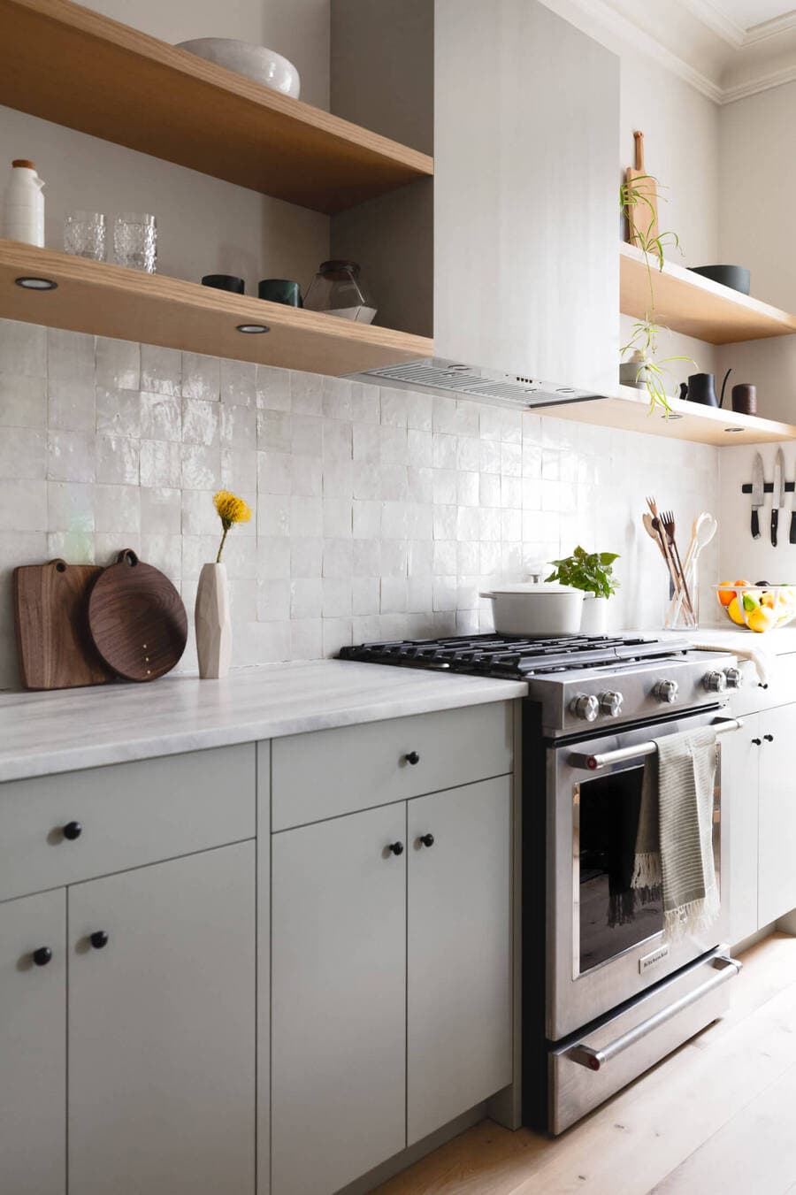 Cocina moderna con muebles color gris, muros de cerámica cuadrada color hueso. Cocina a gas y sobre ella gran campana de acero, combinada con 4 repisas flotantes de madera con luces empotradas en la parte inferior