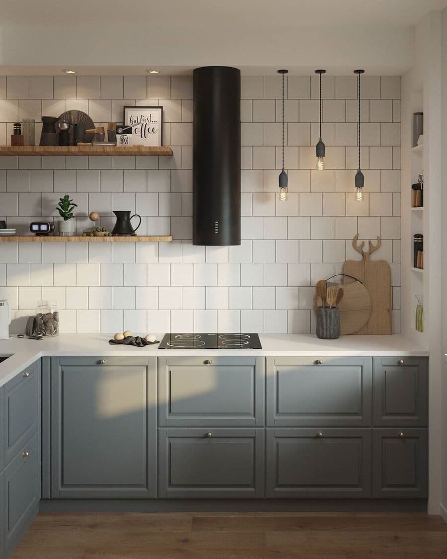 Cocina con muebles de madera color gris y pequeños tiradores. Muros con cerámicas cuadradas blancas. Cocina de vitrocerámica y campana redonda color negro. Tres lámparas colgantes con ampolleta a la vista y repisas flotantes