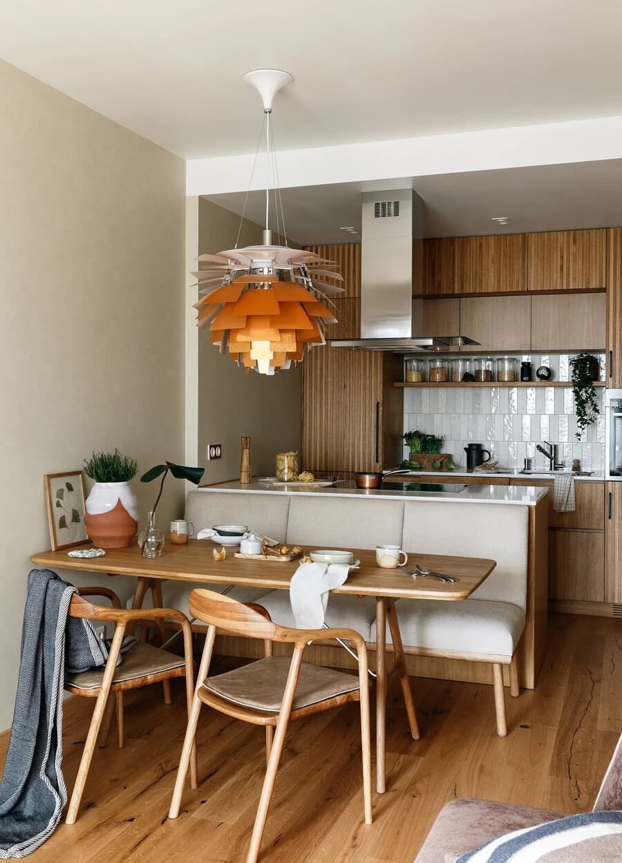 Departamento pequeño con cocina integrada. Muebles de madera con una isla, sobre ella una campana de acero inoxidable. Comedor de madera con dos sillas y una banqueta. Lámpara de techo moderna, tipo "flor"