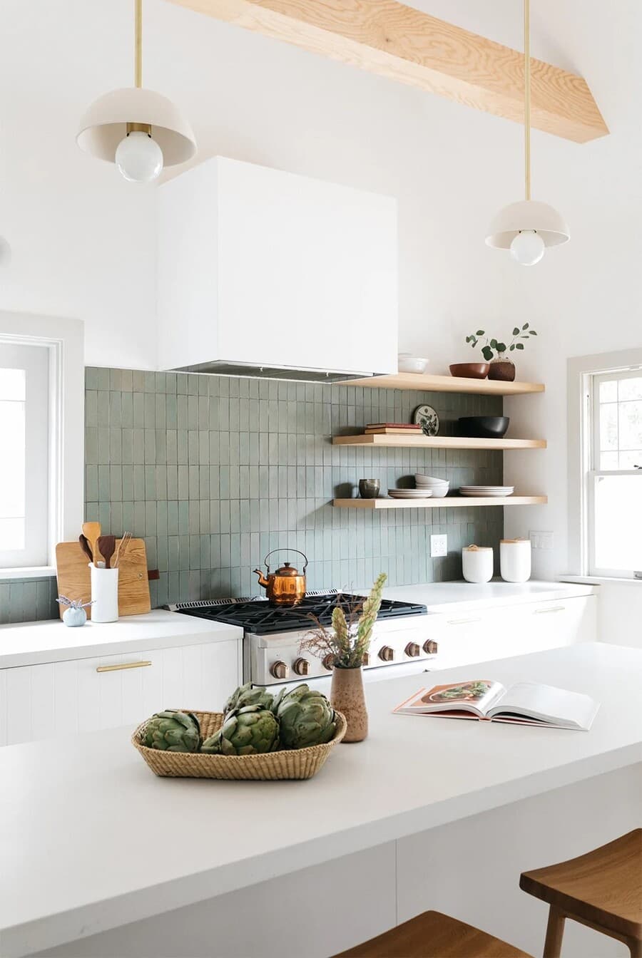 Gran cocina abierta de estilo moderno, muros blancos con media pared con cerámicos gris verdosos. Tres repisas flotante de madera con vajilla. Campana cubierta con revestimiento blanco, sobre una cocina a gas