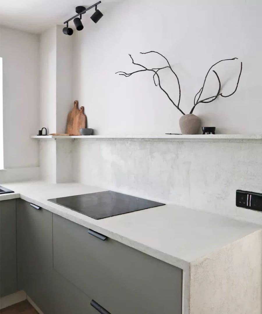 Cocina de estilo minimalista, con cubierta de concreto color gris claro. Muebles con puertas grises y tiradores negros. Muro blanco con una repisa larga, flotante. Sobre ella hay un jarrón de cerámica con ramas secas y una tabla de madera.