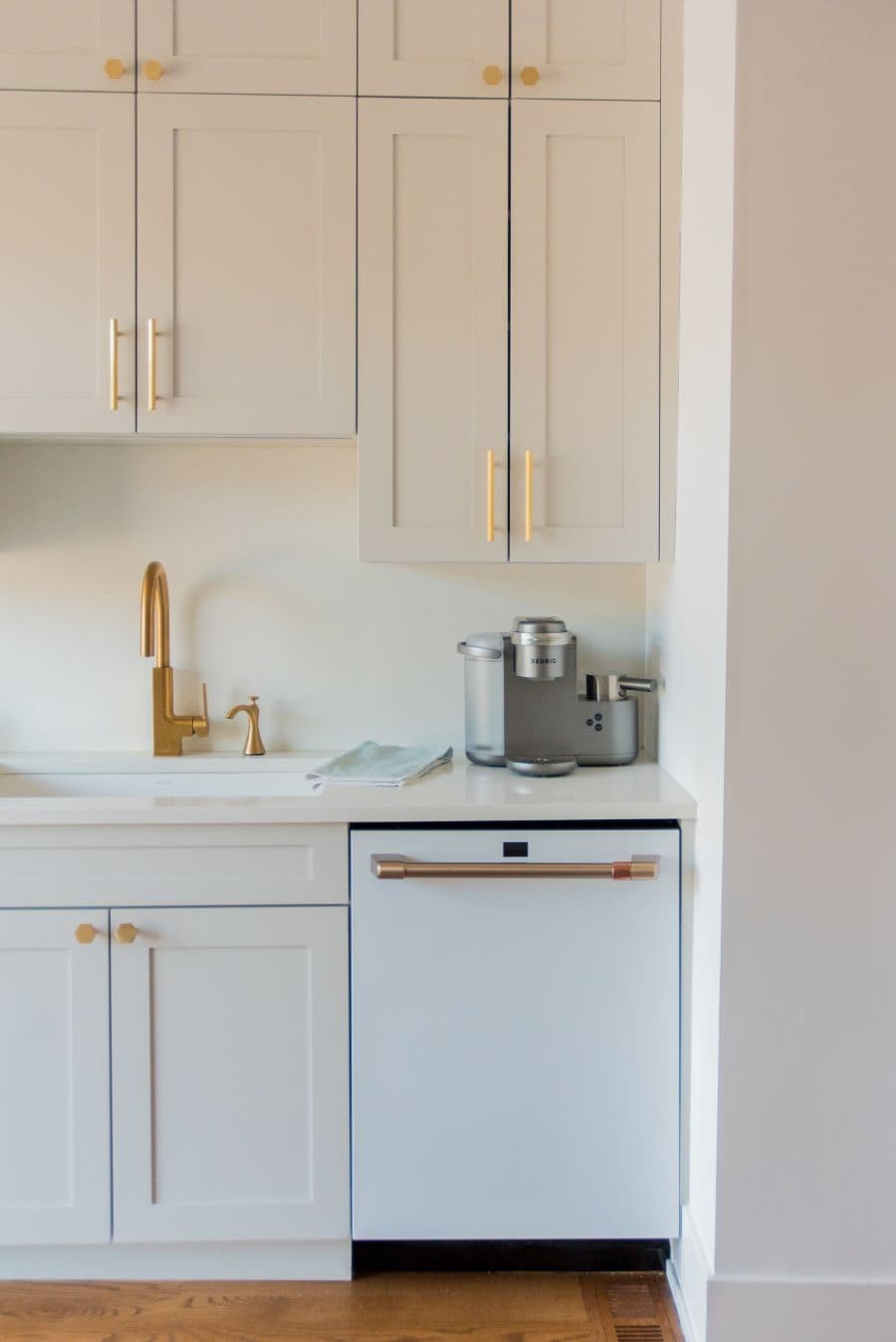 Cocina de estilo clásico, con muebles de madera color gris muy claro y tiradores dorados. Cubierta de cuarzo color beige. Llave de lavaplatos dorada. Sobre el mesón hay una cafetera de estilo moderno. 