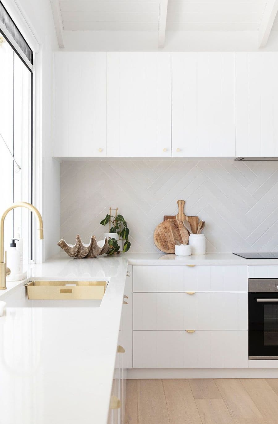 Cocina de estilo moderno, de color blanco, con cubiertas de granito. Llave de lavaplatos de color dorado. Muebles con puertas blancas  y tiradores dorados. Sobre los mesones hay dos tablas de madera, un recipiente de cerámica con cucharas, y una planta.