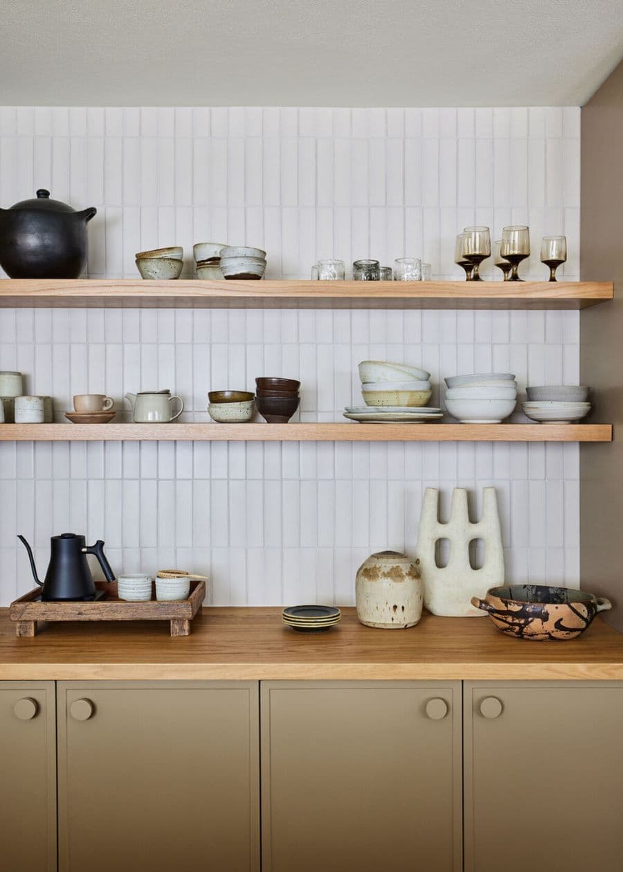 Cocina con cubierta de madrea. Muebles color gris verdoso. Muros con cerámicas rectangulares, de color blanco. Dos repisas flotantes de madera, con vajilla de cerámica gres.