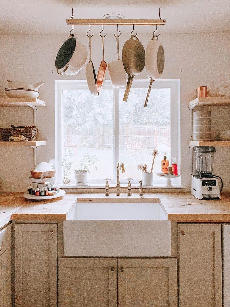 Cocina pequeña con cubierta de madera, pared, muebles y lavaplatos blancos y repisas flotantes de madera. En el techo hay una barra de metal con ganchos donde cuelgan sartenes y ollas.