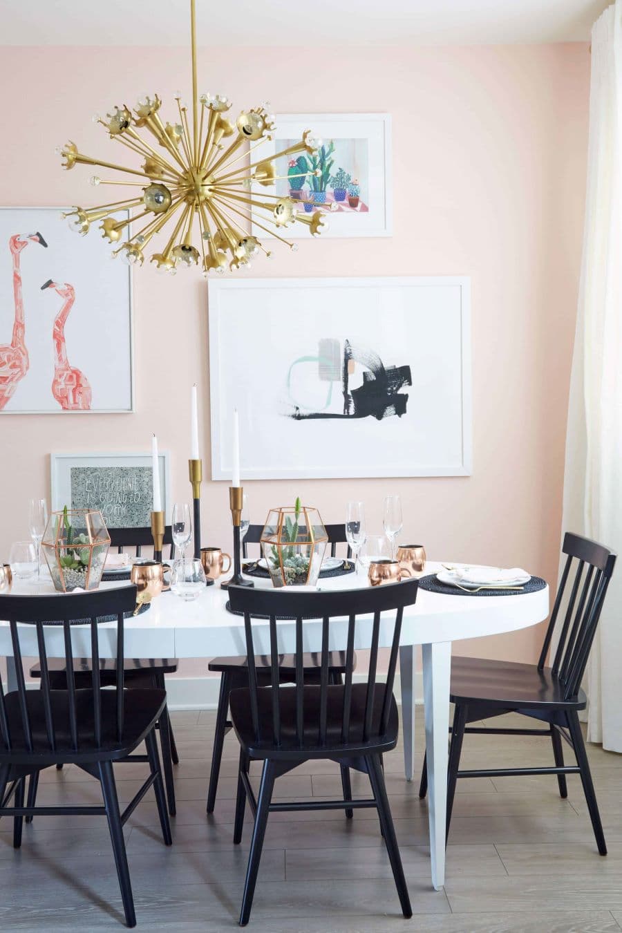Comedor ecléctico, con una mesa blanca y sillas negras. Muros rosa con cuadros y una gran lámpara estilo mid century color dorado.