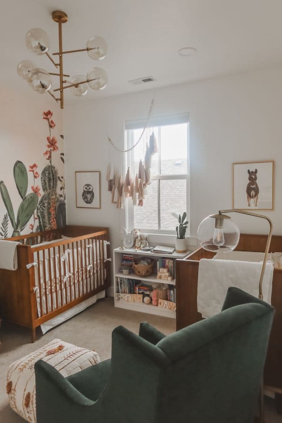 Dormitorio de bebé estilo western. Mural de cactus, cuna de madera oscura. Sitial verde y lámpara de pie con pantalla de vidrio redonda.
