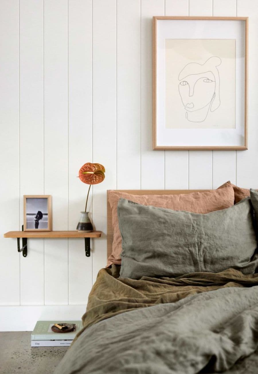 Detalle de las almohadas de una cama en tonos grises y tela lino. Junto a la cama hay un velador flotante de madera, sobre un muro con revestimiento de madera blanca.