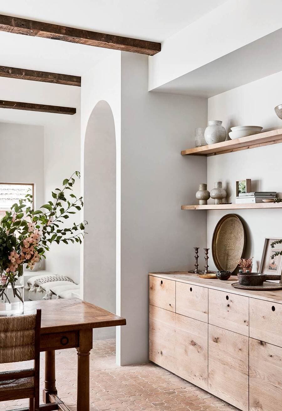 Comedor estilo mediterráneo con dos repisas flotantes de madera sobre muro blanco. En ellas hay adornos y vajilla de cerámica y piezas de artesanía. Bufette de madera con cajones y mesa de comedor de madera