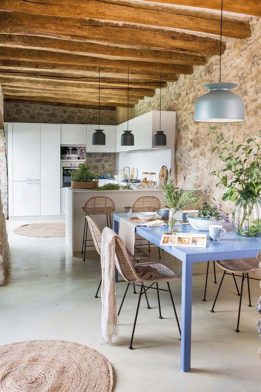 Cocina abierta de estilo mediterráneo, muros de piedra y techo con vigas de madera. Muebles de cocina blancos con isla con tres pisos negros y tres lámparas colgantes. Mesa de comedor azul con sillas de mimbre. Lámpara colgante gris