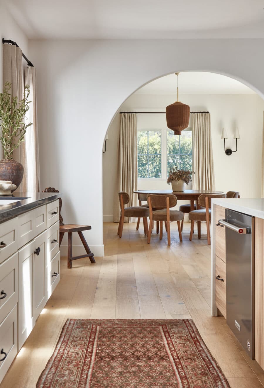 Cocina y comedor mediterráneo, espacio abierto con gran arco central. Muros blancos y piso de madera clara. Muebles de cocina de madera blanca, silla de madera en una esquina. Comedor redondo con seis sillas de tapiz gris