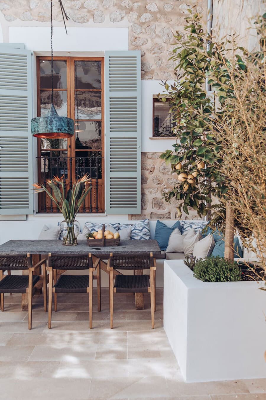 Terraza estilo mediterráneo. Mesa gros con 3  sillas negras y base de madera. Banqueta en L hecha en obra con muchos cojines en tonos azules. Lámpara colgante color azul. Gran ventana con puertas de madera y casa de piedra