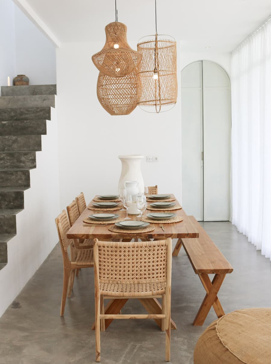 Comedor estilo mediterráneo. Espacio amplio con muros blancos y piso de concreto. Gran mesa de madera, con 5 sillas de fibras naturales, y una banqueta de madera. Desde el techo cuelgan 3 grandes lámparas de mimbre.