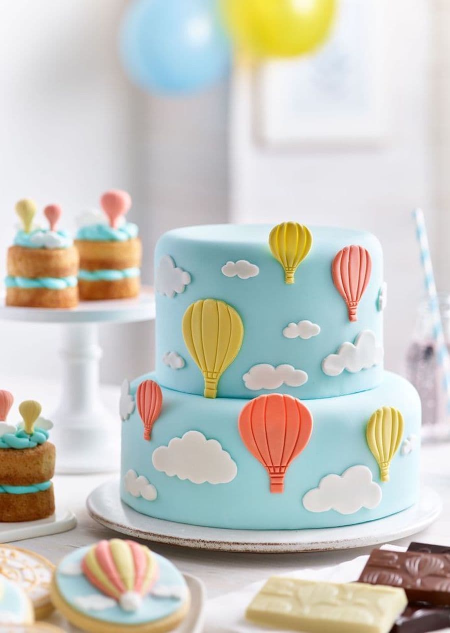 Detalle de una mesa de baby shower con una torta de 2 pisos de nubes y globos aerostáticos amarillos y naranjas, sobre un fondo celeste. 