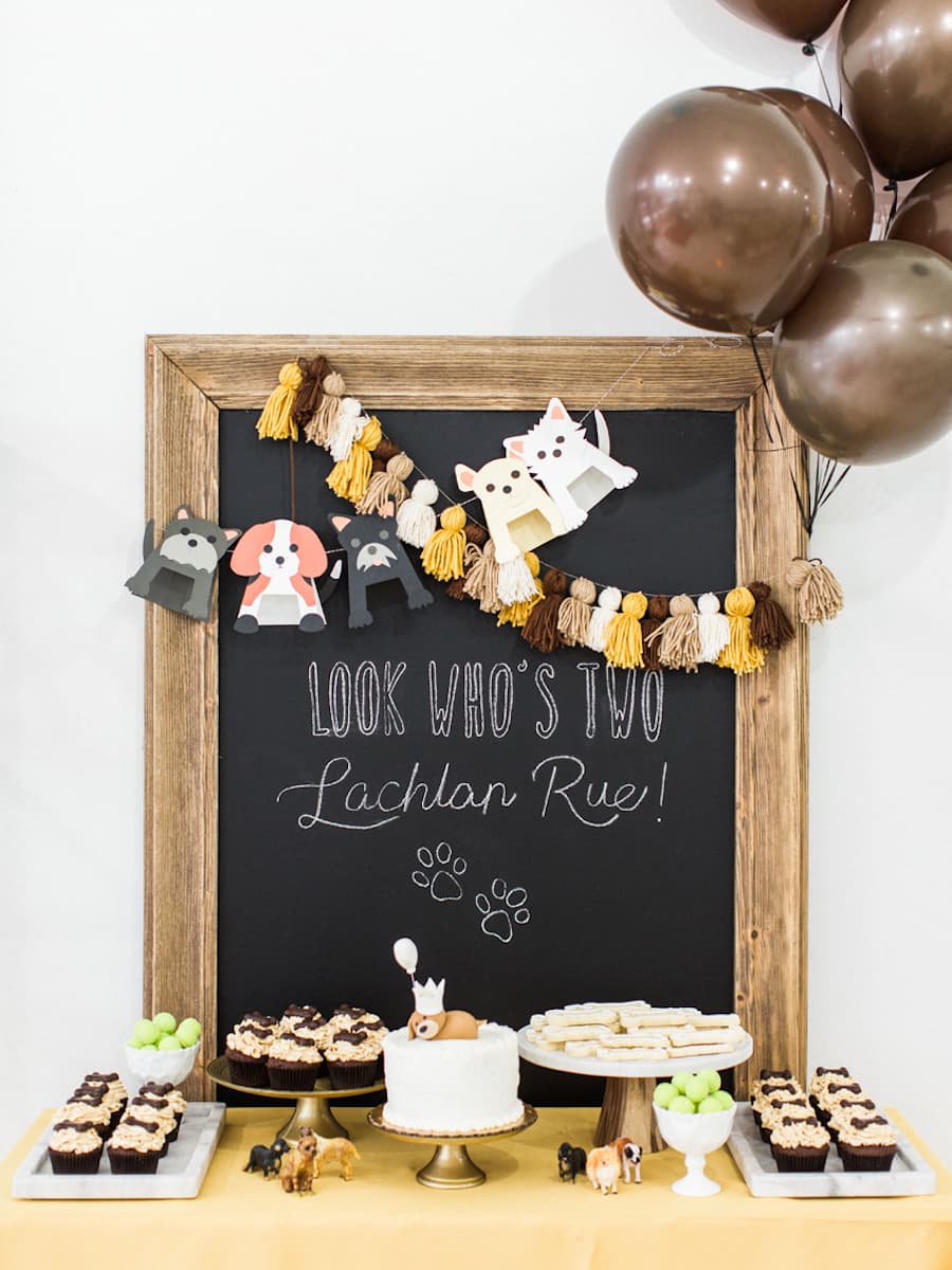 Mesa con distintos sánwiches, cupcakes, dulces y pasteles con motivos de mascotas. Detrás, apoyada sobre una pared, hay una pizara de tiza de marco dorado con una frase en inglés y el nombre del perro de cumpleaños, así como una guirnalda de perros.