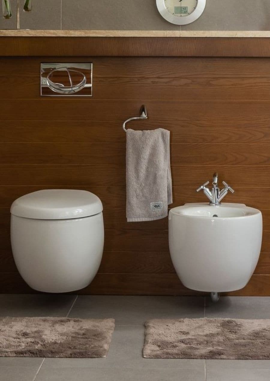 Baño estilo clásico con pared con revestimiento de madera. WC y bidet en set, de forma curva y color blanco. Al medio de ambos hay un colgador con una toalla color gris. Piso de cerámica gris con dos pisos de baño color café.