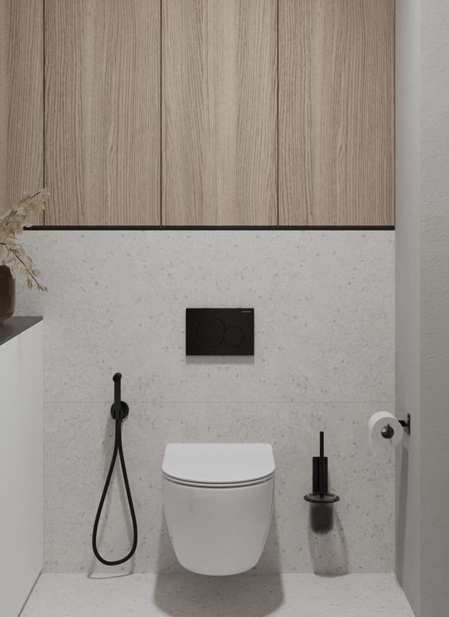 Baño moderno con inodoro de loza blanco y ducha higiénica con manguera negra. Muros de cerámica, estilo terrazo, de color gris claro. En la partes superior hay puertas de madera