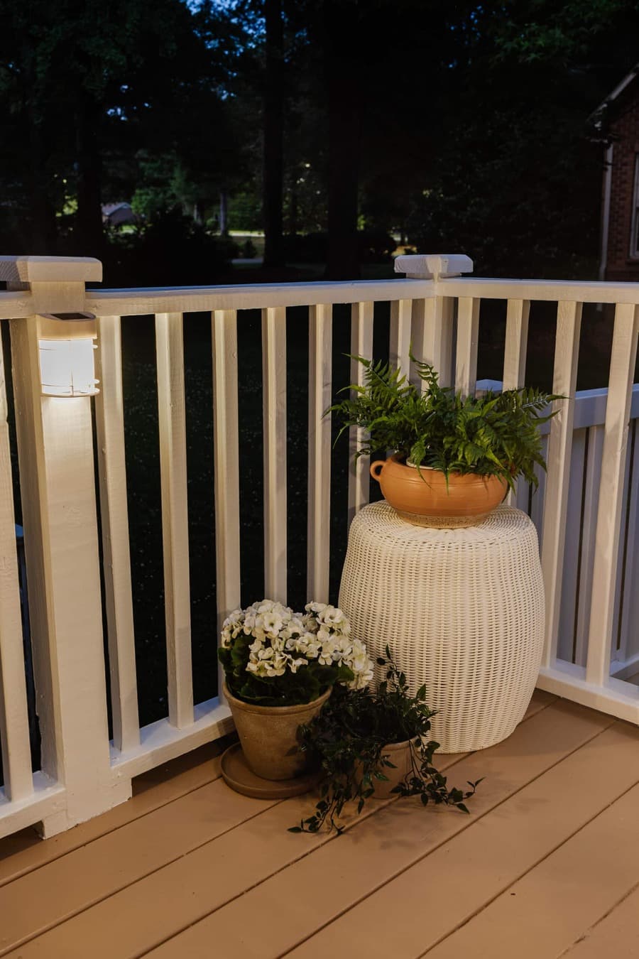 Esquina de una terraza, con una baranda blanca. Uno de los pilares tiene una luz decorativa solar, encendida. En el rincón hay un piso blanco con una maceta con un helecho. A su lado hay otras plantas y flores, también en maceteros.