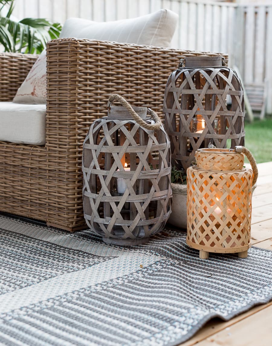 Detalle de una terraza. Sillón de mimbre con cojines blancos, junto a él hay tres fanales de fibras naturales, con velas led dentro. Dos de ellos son grises y uno es café claro. Están sobre una alfombra con líneas en tonos grises.