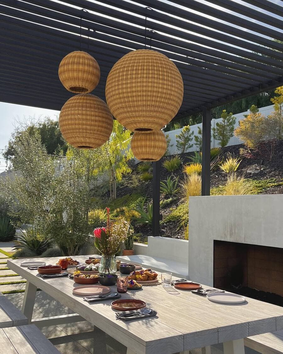 Gran terraza con luces decorativas de mimbre redondas. Son 4 pantallas de diferentes tamaños y colgadas en diferentes alturas, sobre una gran mesa de madera. Hay una parrilla hecha en obra y una estructura metálica negra como techo.