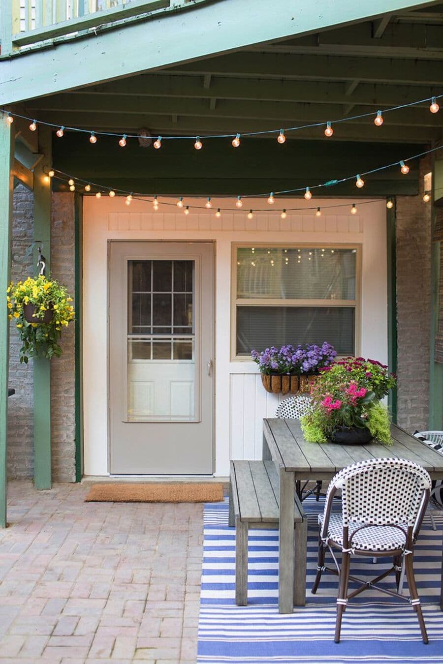 Terraza techada con guirnalda de luces con ampolletas que cruzan el techo. Hay una mesa de madera, tipo picnic, con dos sillas de café estilo francés. Bajo la mesa hay una alfombra en tonos blancos y azules. Al fondo hay una puerta con vidrio y una ventana