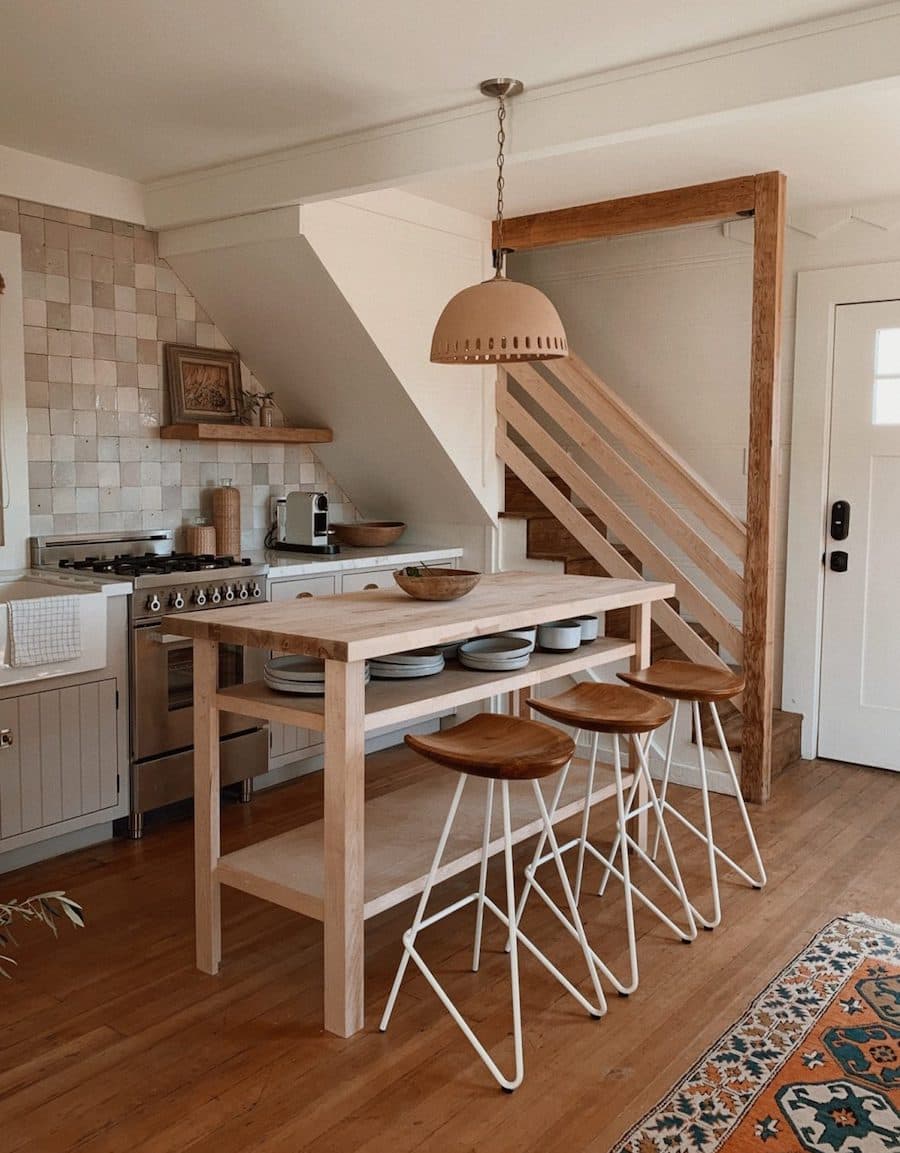 Cocina de estilo rústico con una mesa de madera clara al centro que funciona como isla. Junto a ella hay 3 pisos de bar de patas blancas y asientos de cuero.