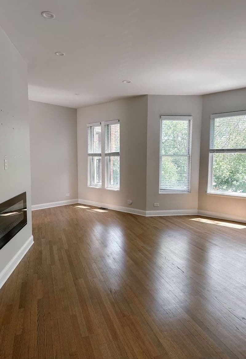 Living vacío, sin muebles, de una casa luminosa. La habitación tiene paredes grises y piso de madera. Tiene una chimenea eléctrica empotrada y varias ventanas que dan a un patio.